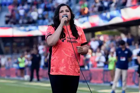 Laura Bravo singing the national anthem at a stadium in a coral team jersey