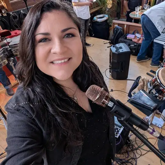 Laura Bravo smiling beside her microphone at a venue, with the band's gear set up behind her